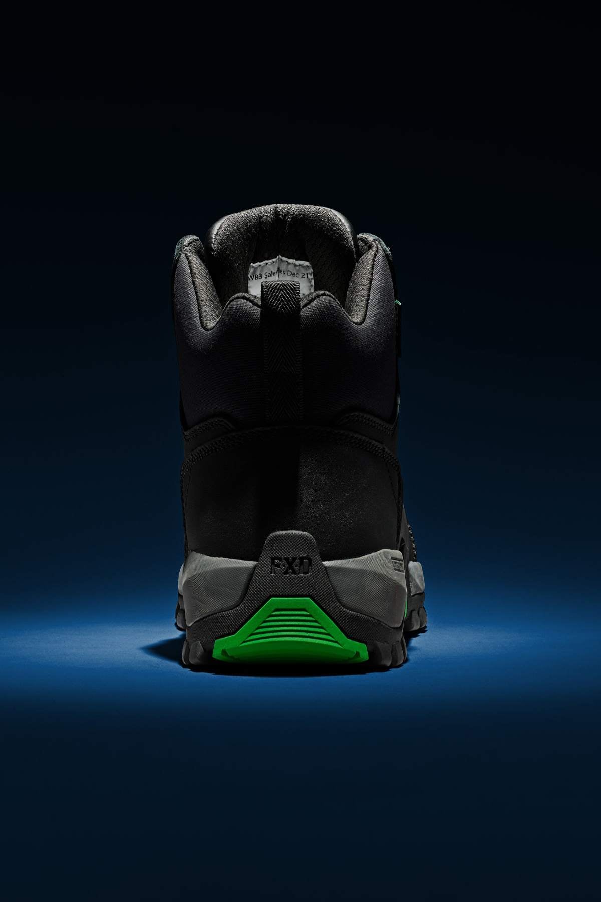 The FXD WB-3 Work Boot, displayed from the rear, rests on a dark blue surface against a black backdrop. It is made from premium leather and showcases a green accent on the sole, embodying a rugged design suitable for outdoor use.