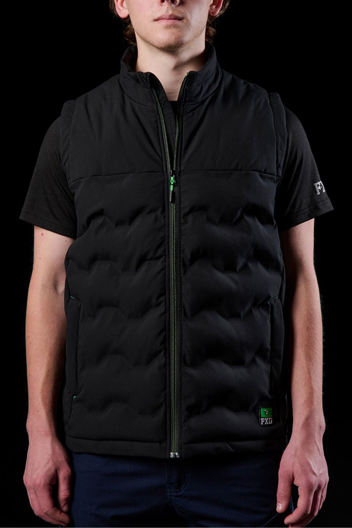 Wearing an FXD WO-6 Insulated Puffer Vest with water repellent fabric over a black short sleeve shirt, a person stands against a black background. The vest has a subtle logo on the lower left, and their face is partly visible.