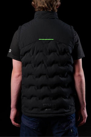 A person is seen from behind wearing the FXD WO-6 Insulated Puffer Vest over a black short-sleeve shirt, set against a black background. The vest features performance fabric and a small green horizontal line near the top.