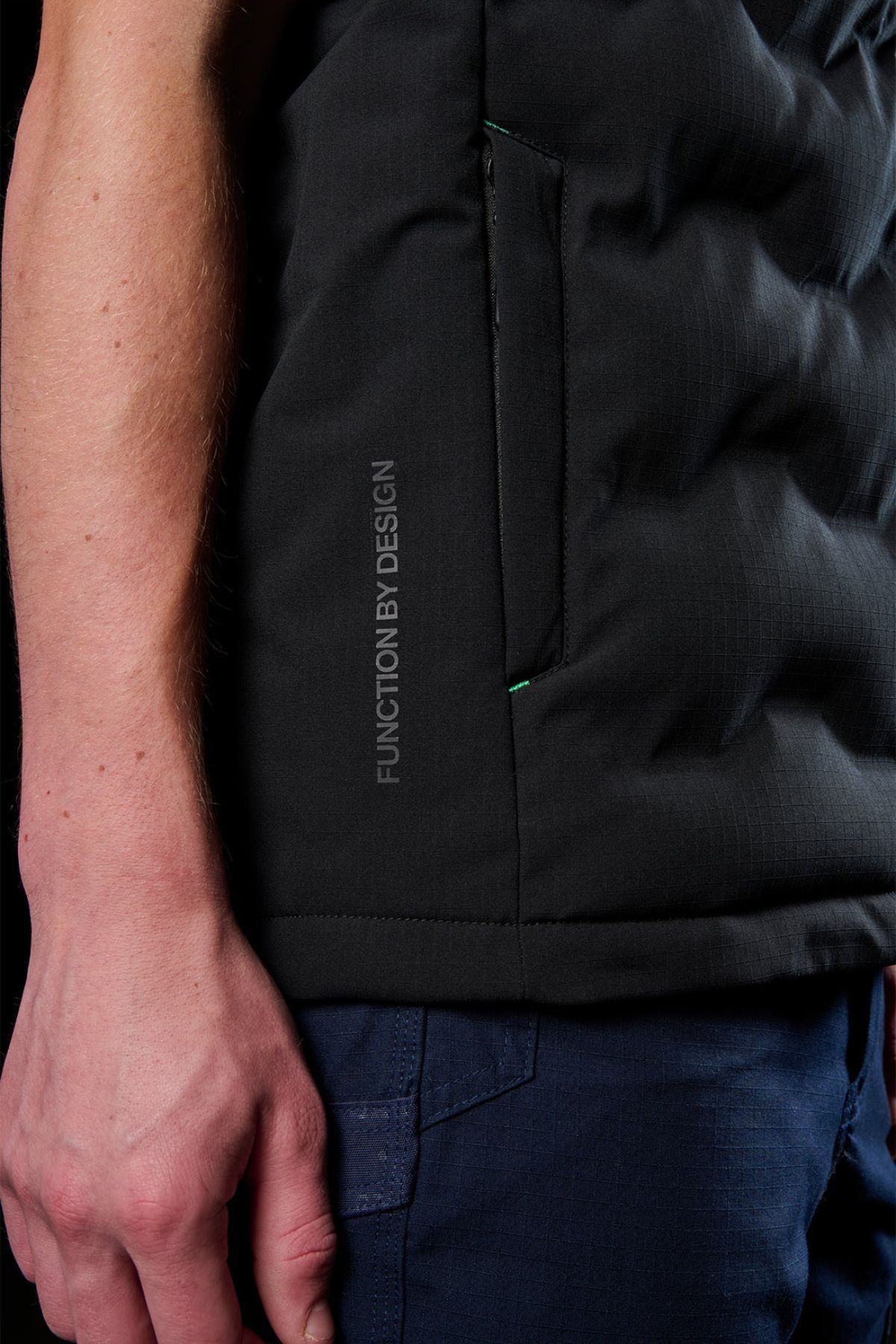 A close-up of an arm and hand wearing a black FXD WO-6 Insulated Puffer Vest with "FUNCTION BY DESIGN" near the side pocket, paired with dark blue pants, against a dark background.