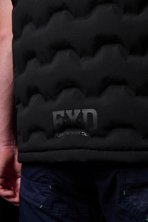 Close-up of a person wearing the FXD WO-6 Insulated Puffer Vest in black with a textured finish and dark blue pants. The FXD logo and "FUNCTION BY DESIGN" are visible near the bottom. Only part of their arm is shown.