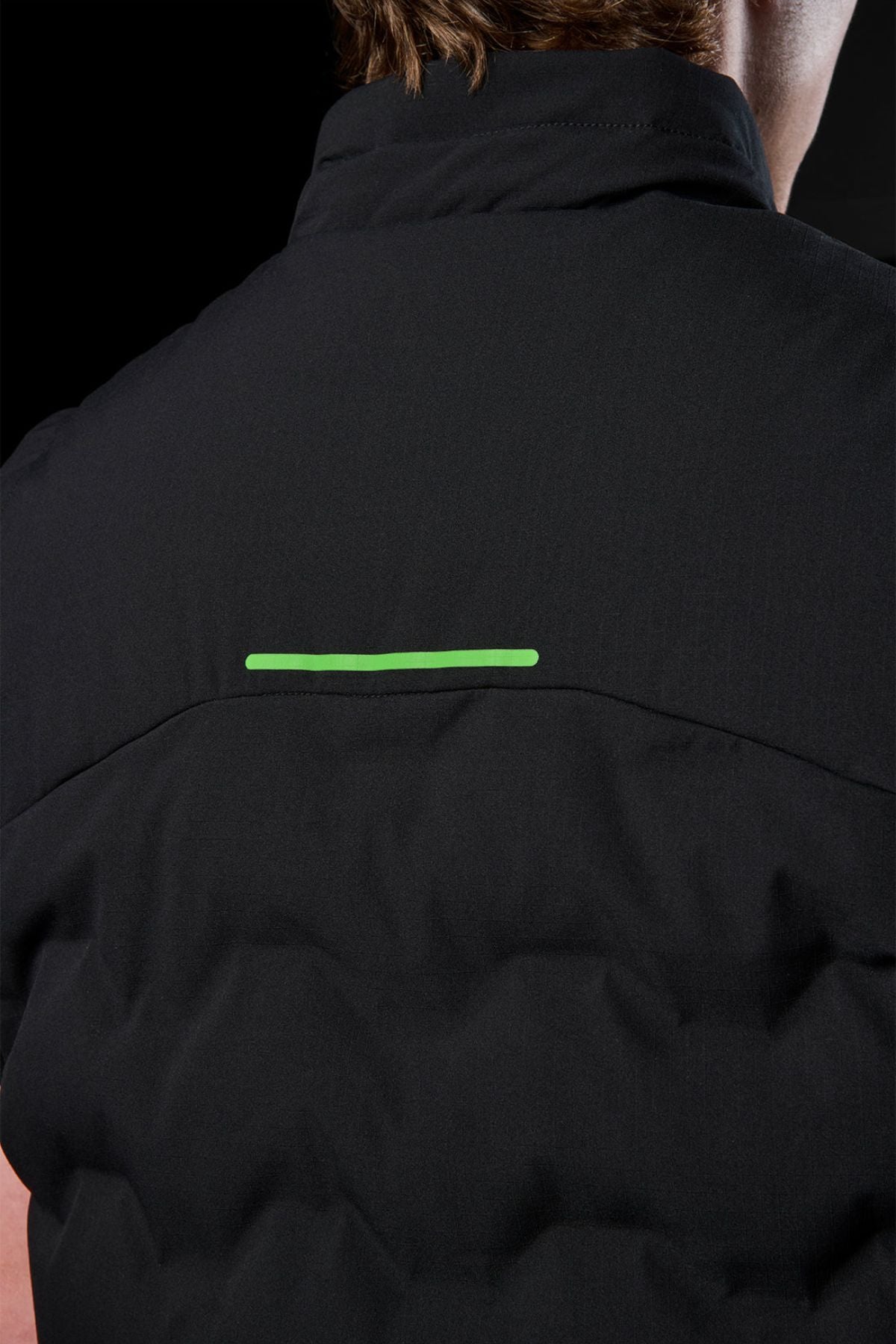 Close-up of a person's upper back wearing the FXD WO-6 Insulated Puffer Vest, featuring black performance fabric and a horizontal neon green reflective strip across the shoulders, set against a black background.