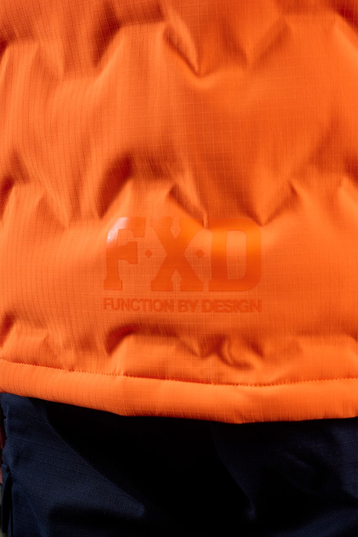 Close-up of the FXD WO-6 Hi Vis Insulated Puffer Vest in orange with "FXD" and "Function by Design" printed on it, shown above a section of dark-colored material.