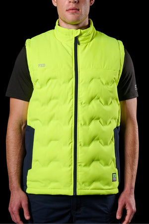 A person wearing the FXD WO-6 Hi Vis Insulated Puffer Vest, a bright yellow sleeveless puffer with black side panels and zipper, over a black short-sleeve shirt, stands against a black background.