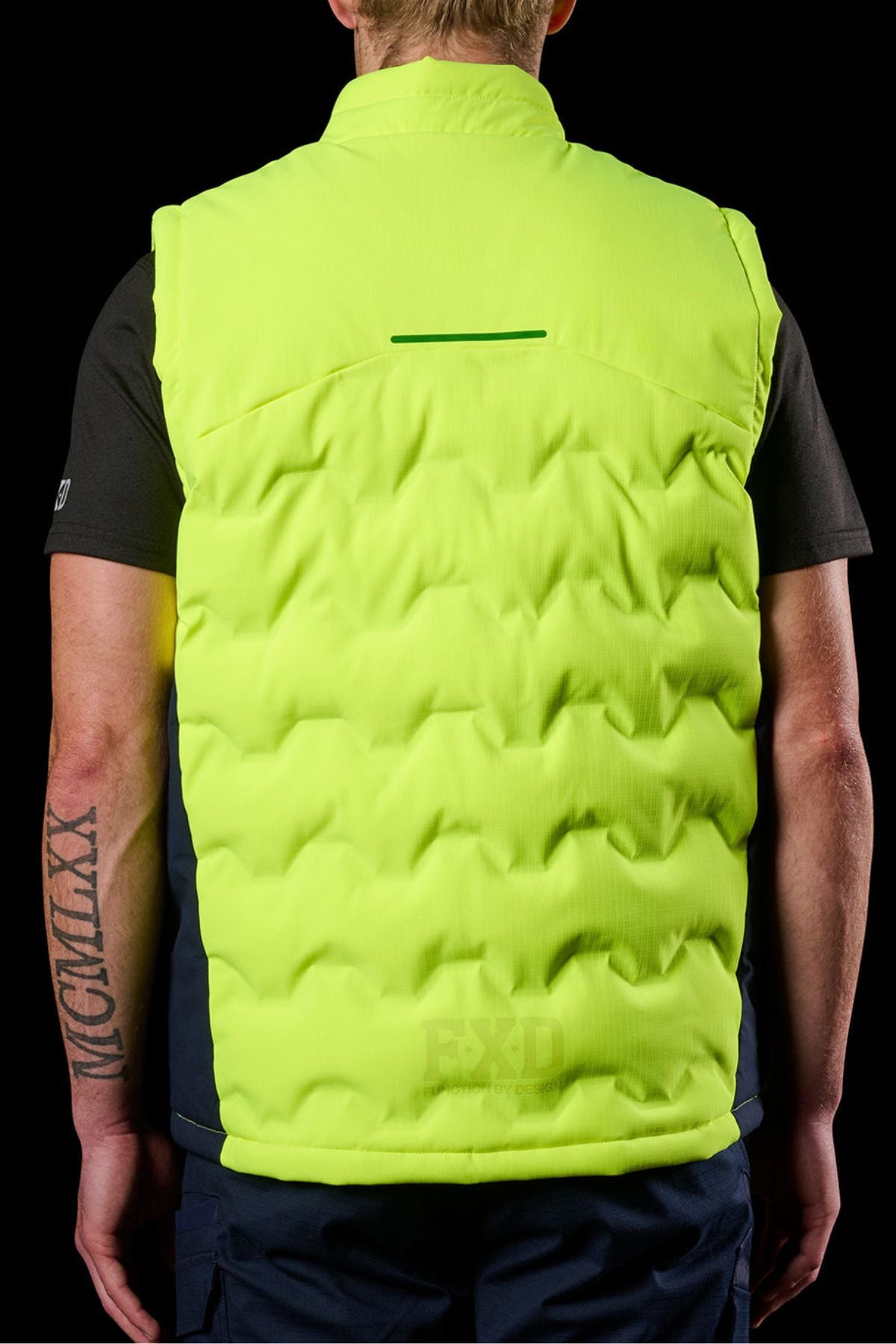 A person is shown from the back wearing the FXD WO-6 Hi Vis Insulated Puffer Vest over a black shirt, with arm tattoos and short blonde hair standing out against a black background.