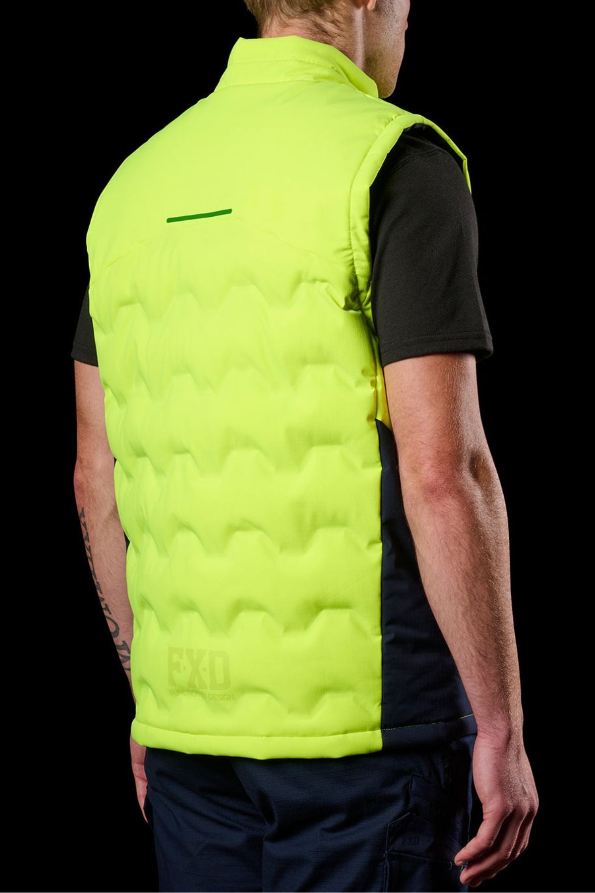 A person seen from behind, wearing the FXD WO-6 Hi Vis Insulated Puffer Vest over a black short-sleeve shirt, against a black background.
