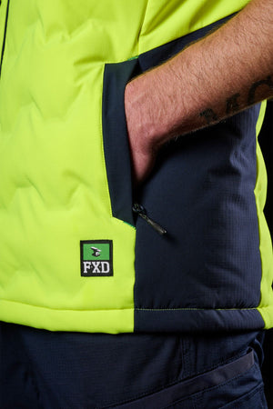 A close-up of a tattooed hand in the pocket of a vibrant yellow and navy FXD WO-6 Hi Vis Insulated Puffer Vest, with the FXD logo visible near the pocket.