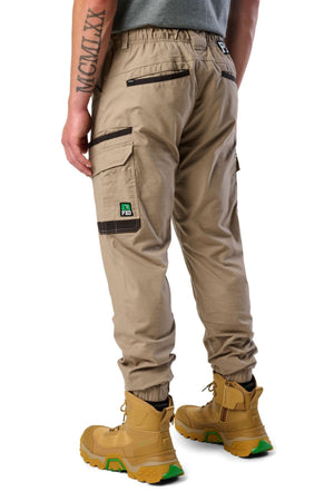 A person shown from the back wears FXD WP-12C stretch ripstop work pants with black zipper pockets and a green and black "FXD PRO" patch, yellow work boots with green soles, and a grey shirt.
