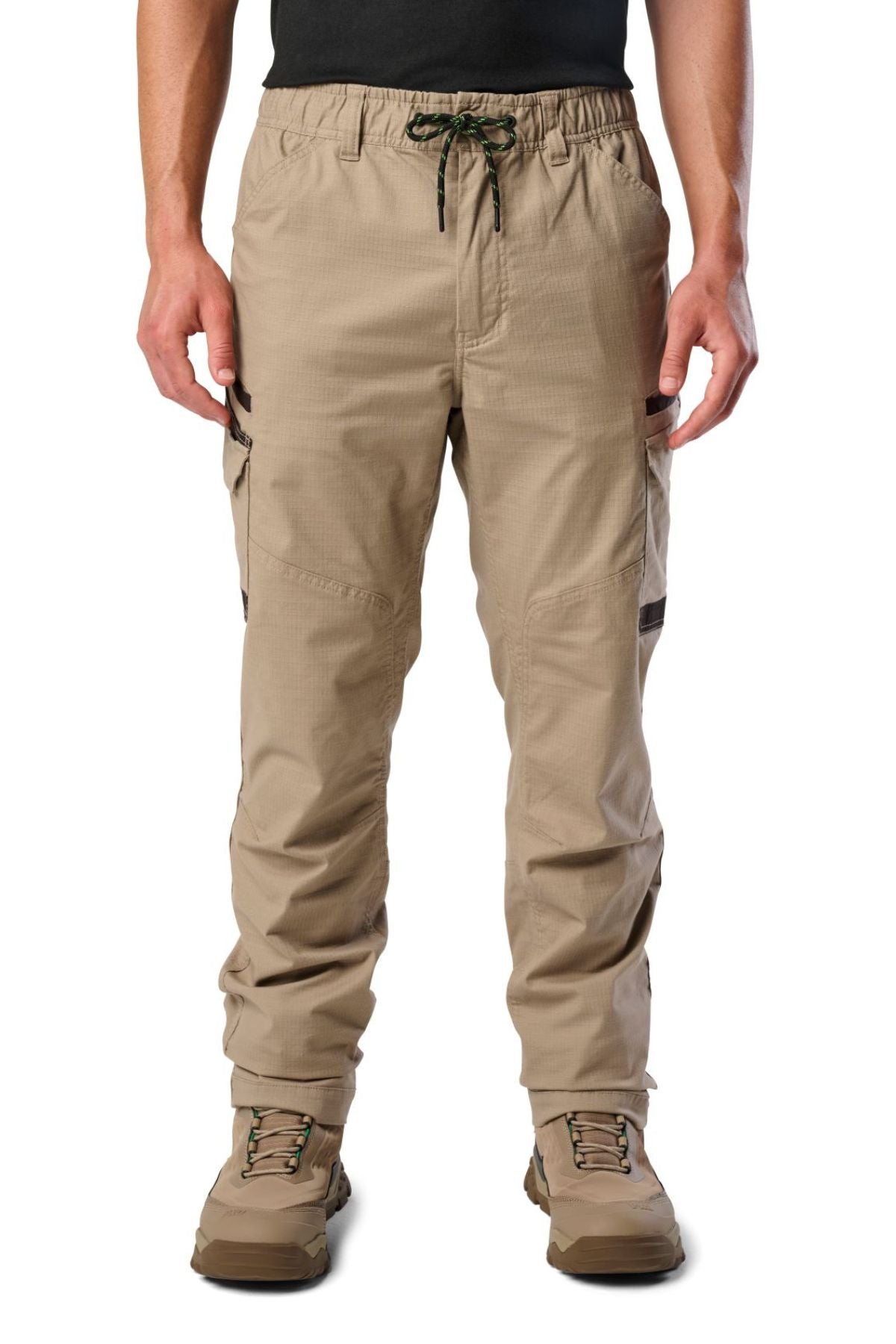 A person stands against a white background wearing FXD WP-12 tan 2-way stretch ripstop pants with utility pockets and a drawstring waist, paired with light brown hiking boots and a black shirt.