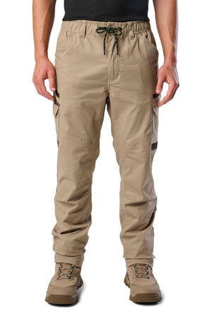 A person stands against a white background wearing FXD WP-12 tan 2-way stretch ripstop pants with utility pockets and a drawstring waist, paired with light brown hiking boots and a black shirt.