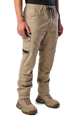 The lower half of a person is shown wearing FXD WP-12 tan 2-way stretch ripstop utility pants with an elastic waistband, drawstring, and pockets, paired with tan hiking boots featuring green accents.