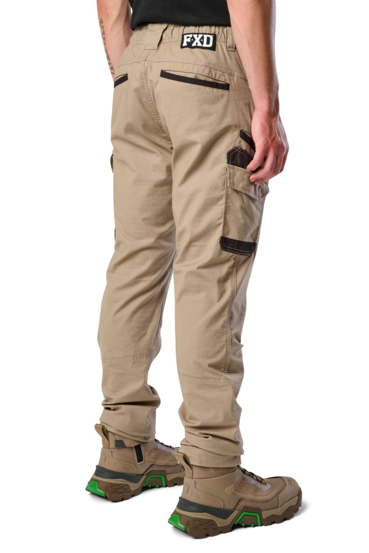 A person models FXD WP-12 tan 2-way stretch ripstop pants with utility pockets and brown details, paired with tan work boots with green soles, standing on a white background facing away from the camera.