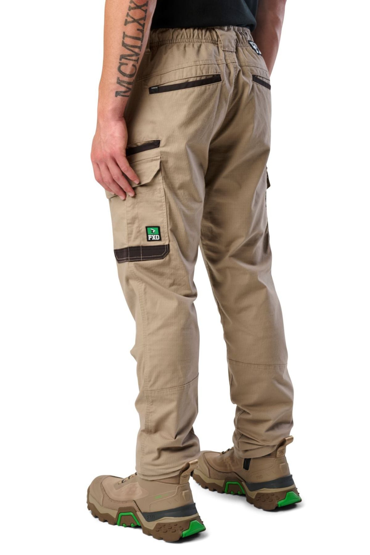 A person wearing FXD WP-12 tan 2-way stretch ripstop pants with black trim, utility pockets, tan work boots with green accents, and a black shirt stands facing away. A tattoo is visible on their left forearm.