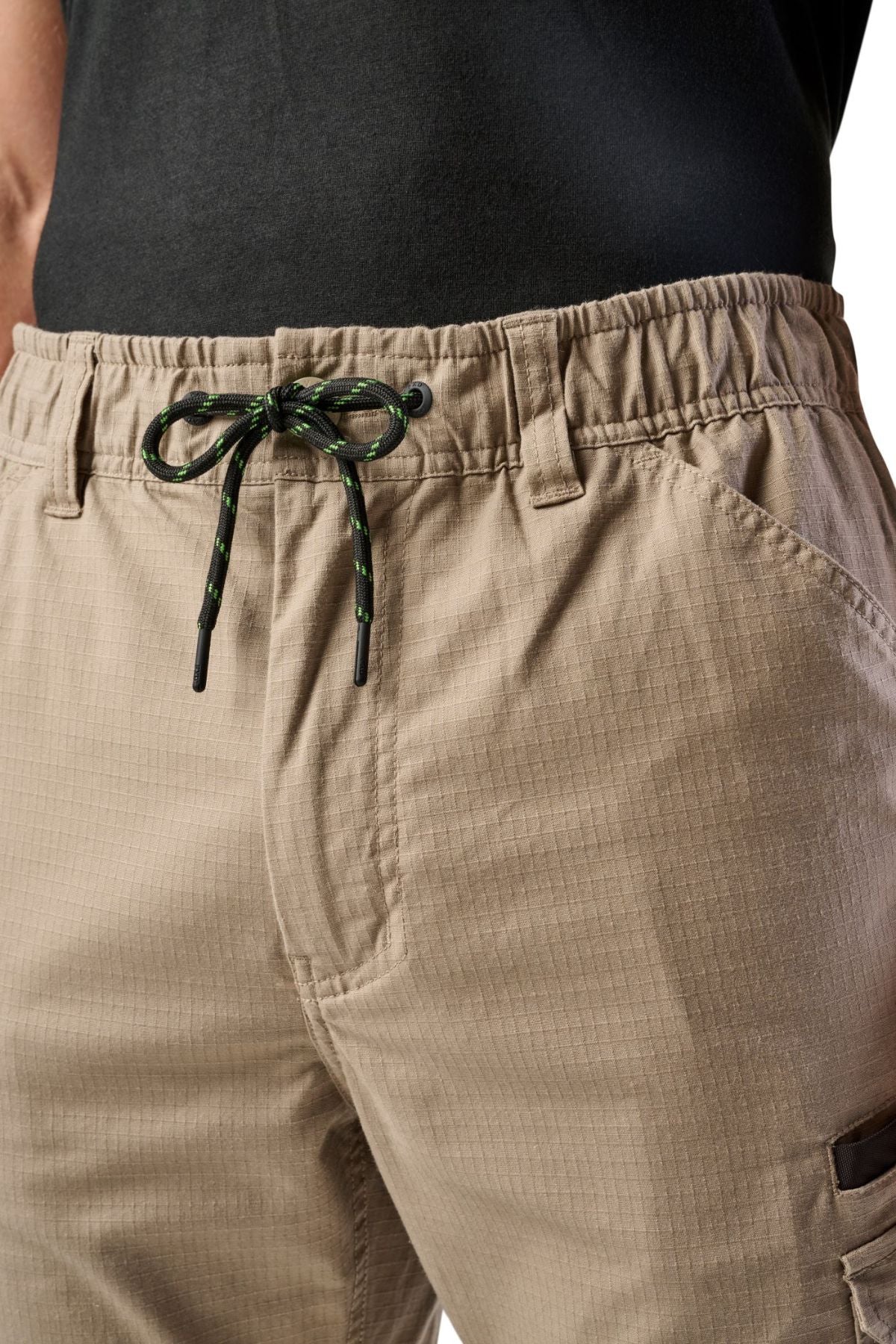 Close-up of a person wearing FXD WP-12 beige 2-way stretch ripstop work pants with utility pockets, a black shirt, and black drawstring with green accents. The elastic waistband enhances comfort and durability.