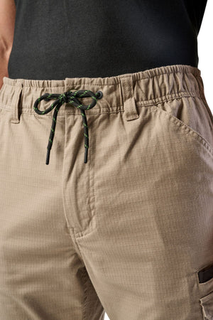 Close-up of a person wearing FXD WP-12 beige 2-way stretch ripstop work pants with utility pockets, a black shirt, and black drawstring with green accents. The elastic waistband enhances comfort and durability.