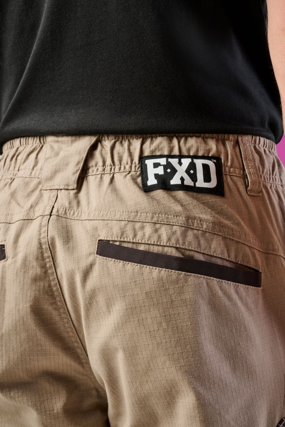 Close-up of a person wearing FXD WP-12 beige durable work pants with utility pockets and a black FXD logo patch above the back pocket, paired with a slightly tucked-in black shirt.