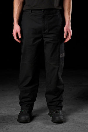 A person stands facing forward, wearing black FXD WP-3L Loose Fit Stretch Canvas Work Pants, a black shirt, and black shoes, with hands relaxed by their sides against a dark background.