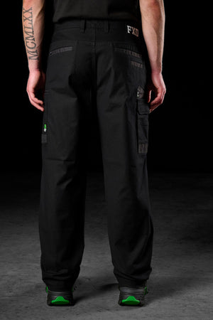 A person stands with their back to the camera, wearing FXD WP-3L Loose Fit Stretch Canvas Work Pants and green-accented black shoes. A Roman numeral tattoo is visible on their right arm against a dark background.