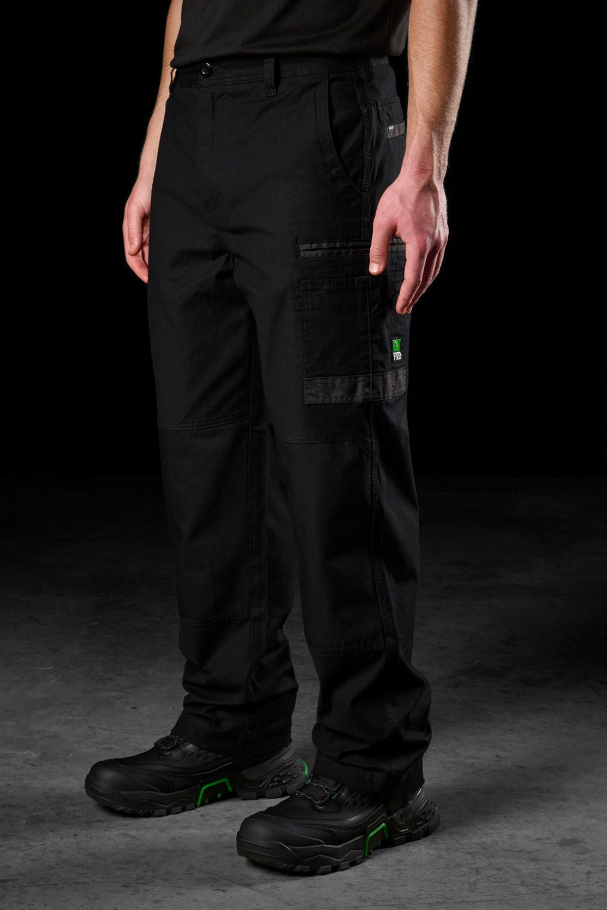 A person stands against a dark background, showing only their lower torso and legs in FXD WP-3L Loose Fit Stretch Canvas Work Pants and black work boots; their upper body and face are not visible.