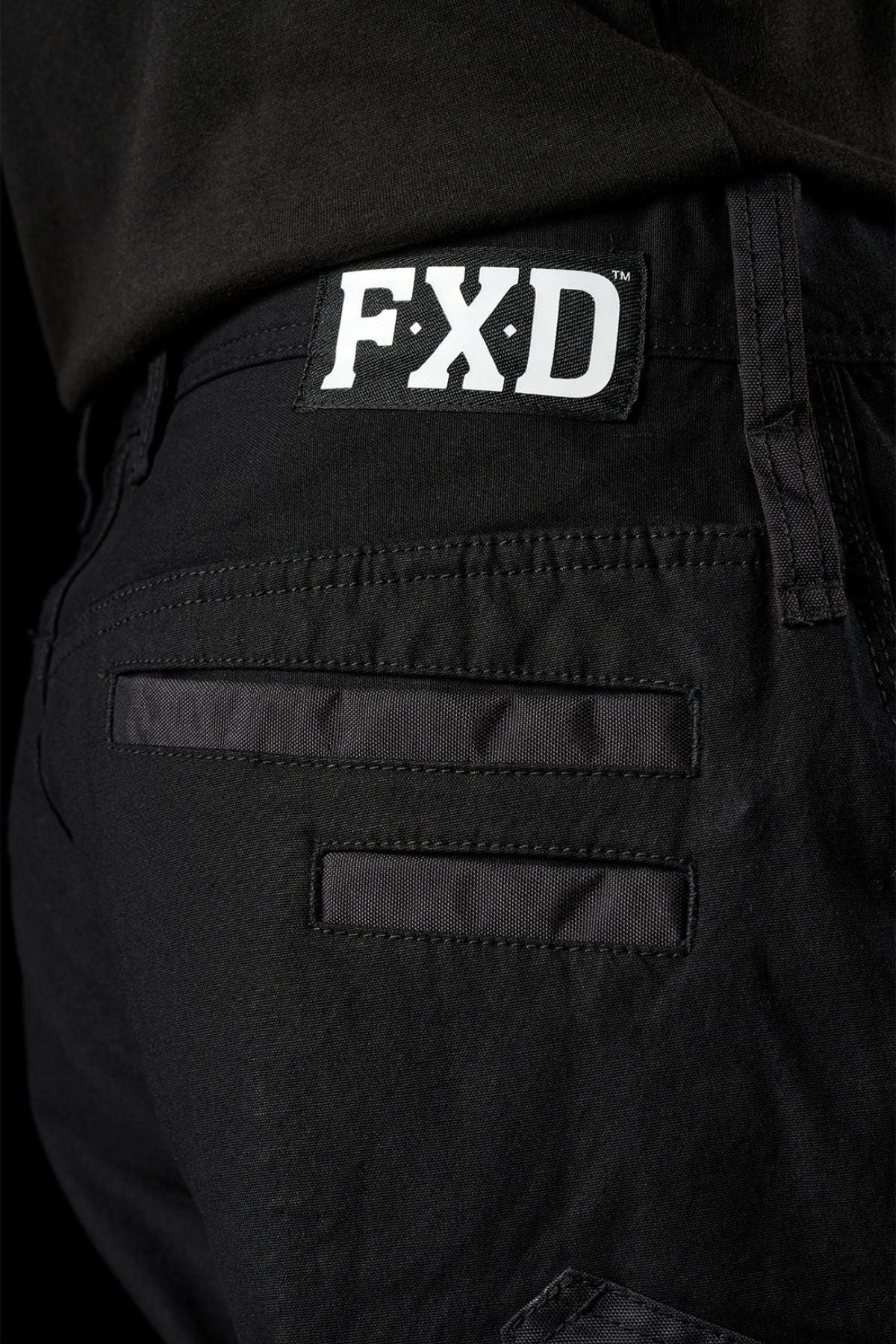Close-up of the back of FXD WP-3L Loose Fit Stretch Canvas Work Pants, showing two horizontal pockets and a white "FXD" logo label above the right one. The durable pants are paired with a black shirt.
