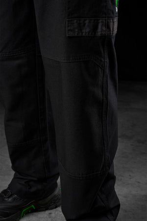 Close-up of a person wearing FXD WP-3L Loose Fit Stretch Canvas Work Pants and black shoes with green accents, standing on a grey concrete floor against a dark background.