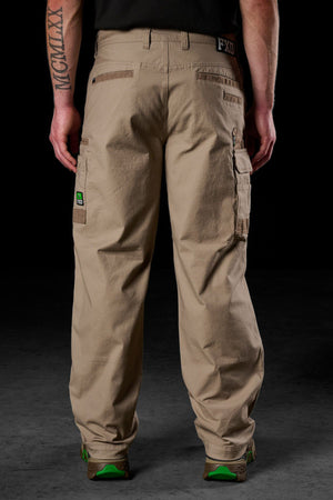 A person faces away, wearing FXD WP-3L Loose Fit Stretch Canvas Work Pants with multiple pockets and green-accented brown shoes. A Roman numeral tattoo is visible on their left forearm against a dark background.
