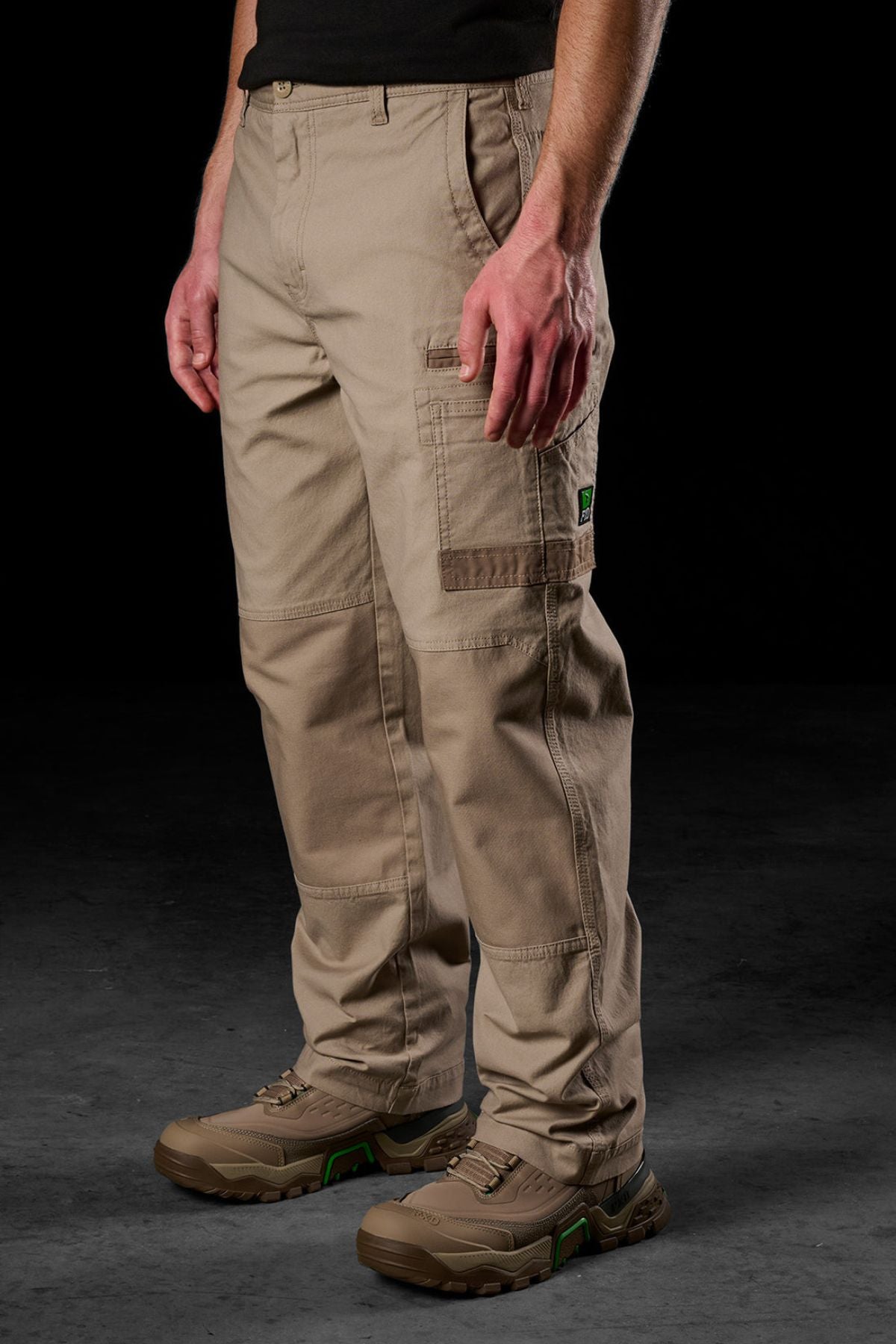 A person wearing tan FXD WP-3L Loose Fit Stretch Canvas Work Pants, a black shirt, and brown work shoes stands against a dark background. The pants offer multiple pockets and reinforced stitching for durability.