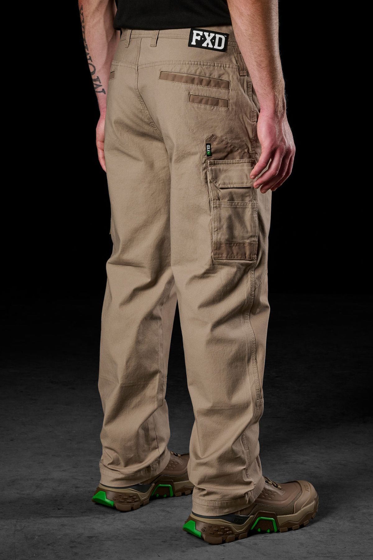 A person in tan FXD WP-3L Loose Fit Stretch Canvas Work Pants with multiple pockets, brown and green boots, and a black shirt with "FXD" on the waistband stands on a dark surface.