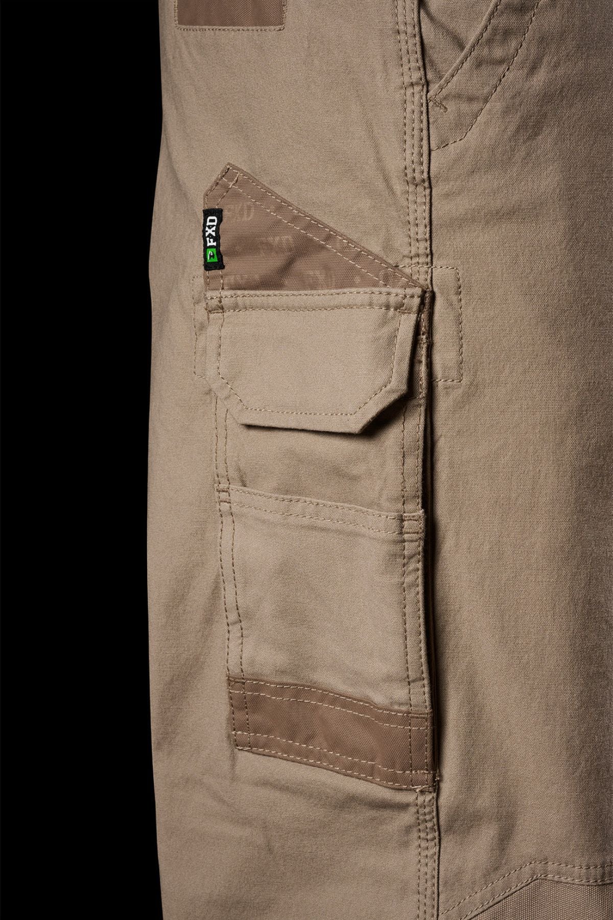 Close-up of a cargo pocket on FXD WP-3L Loose Fit Stretch Canvas Work Pants, showing detailed stitching, a flap with black and green tag, and reinforced edges against a solid black background.