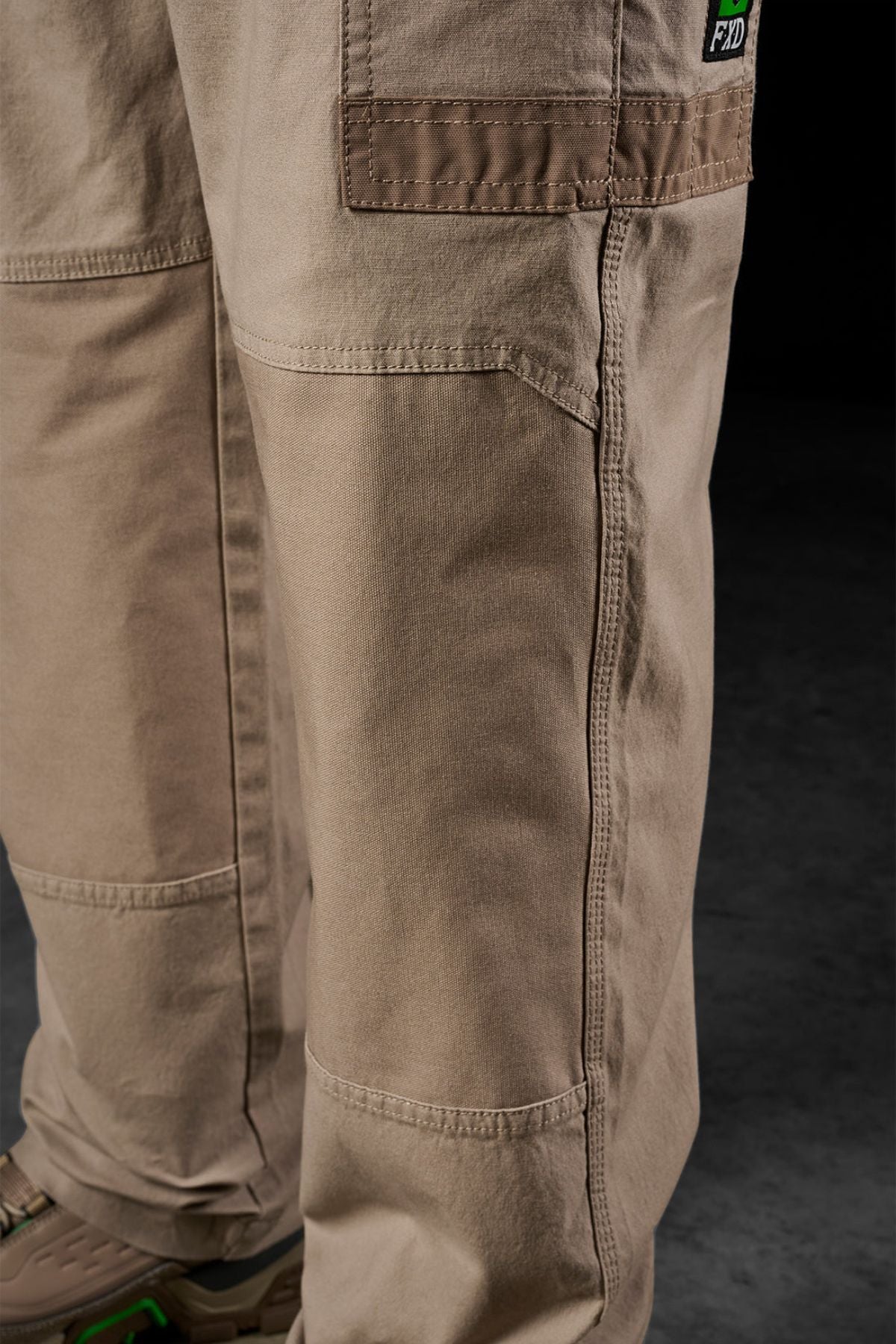 Close-up of a person wearing FXD WP-3L Loose Fit Stretch Canvas Work Pants in light brown with brown shoes featuring green accents, standing on a dark textured floor—showcasing the pants' durable fabric and detailed stitching.