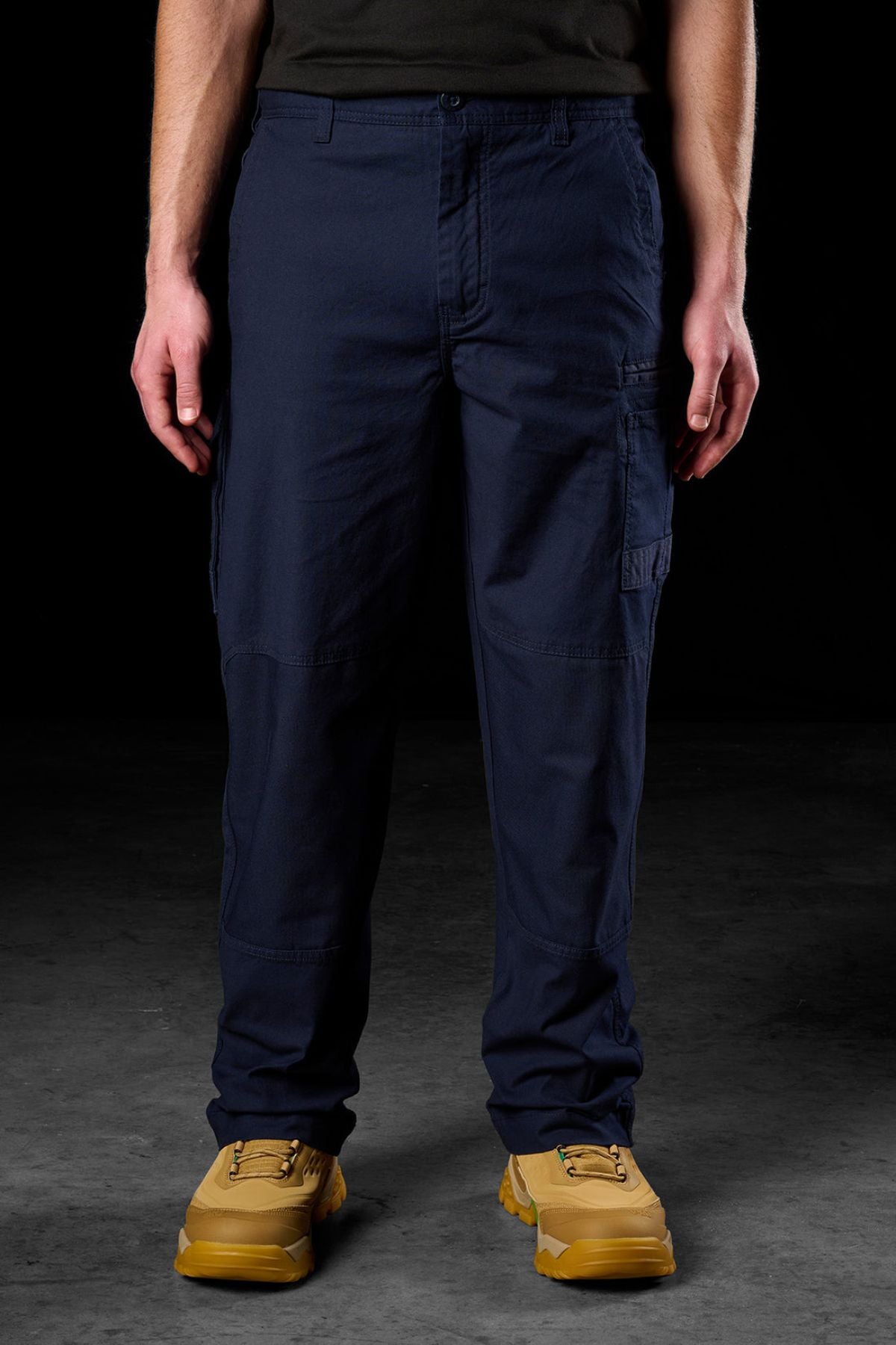 Someone wearing FXD WP-3L Loose Fit Stretch Canvas Work Pants in navy, a black shirt, and tan work boots stands on a dark textured floor with arms relaxed at their sides.