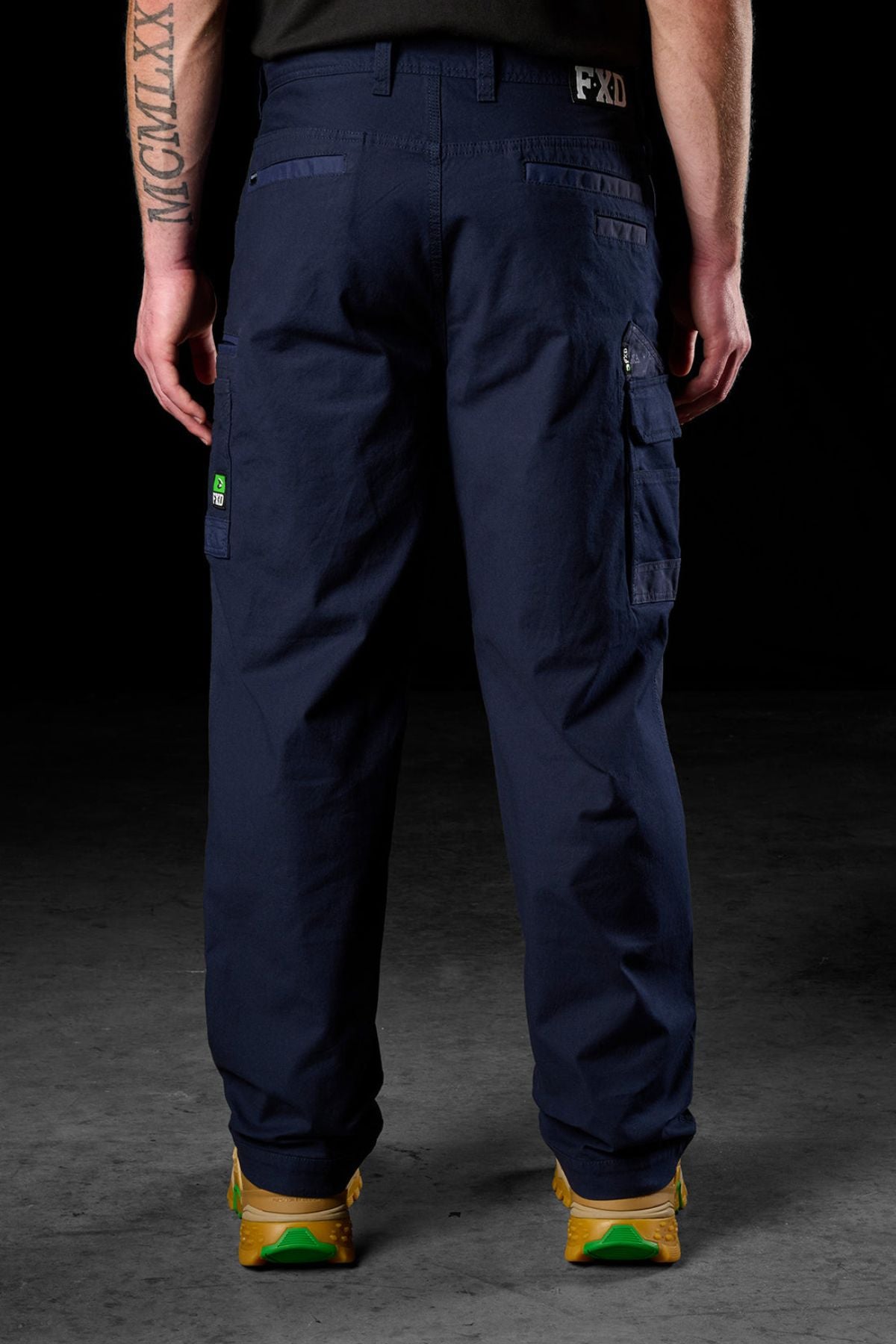 A person faces away from the camera in FXD WP-3L Loose Fit Stretch Canvas Work Pants (navy blue), paired with yellow-green shoes. Tattooed arms are visible against a dark background.
