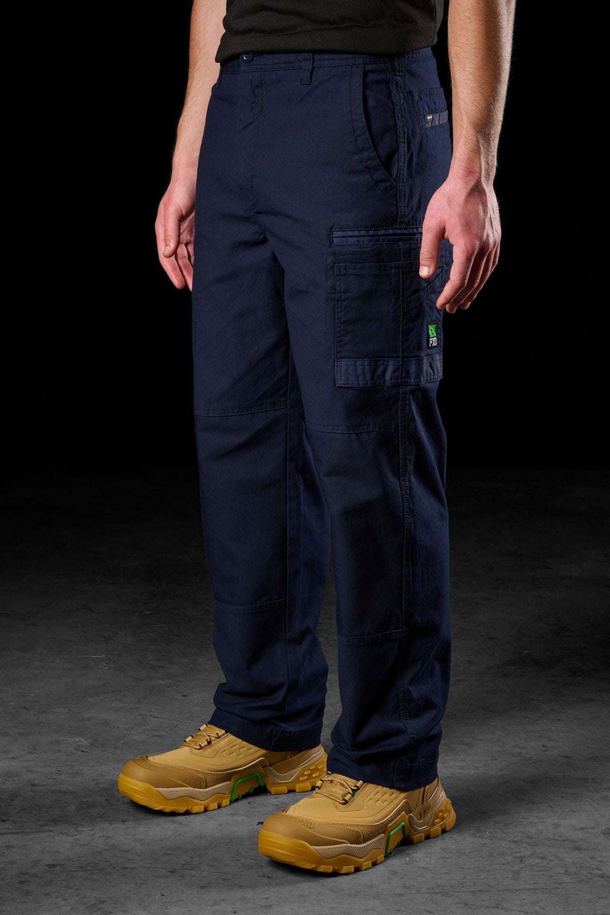 A person stands against a dark background wearing FXD WP-3L Loose Fit Stretch Canvas Work Pants in navy, featuring stretch fabric and multiple pockets, including one with a green tag. Tan work boots complete the outfit; hands rest at their sides.