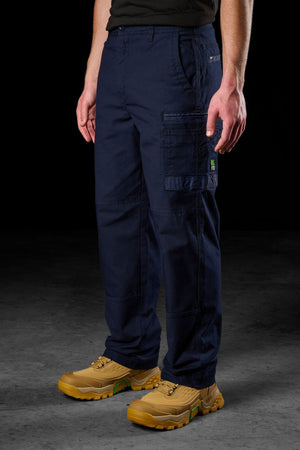 A person stands against a dark background wearing FXD WP-3L Loose Fit Stretch Canvas Work Pants in navy, featuring stretch fabric and multiple pockets, including one with a green tag. Tan work boots complete the outfit; hands rest at their sides.