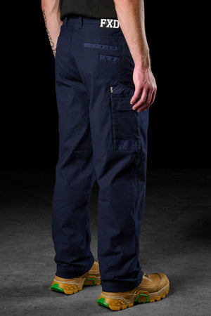 A person stands facing away against a dark background, wearing FXD WP-3L Loose Fit Stretch Canvas Work Pants with multiple pockets and tan work boots with green accents. The FXD logo is visible on the waistband.