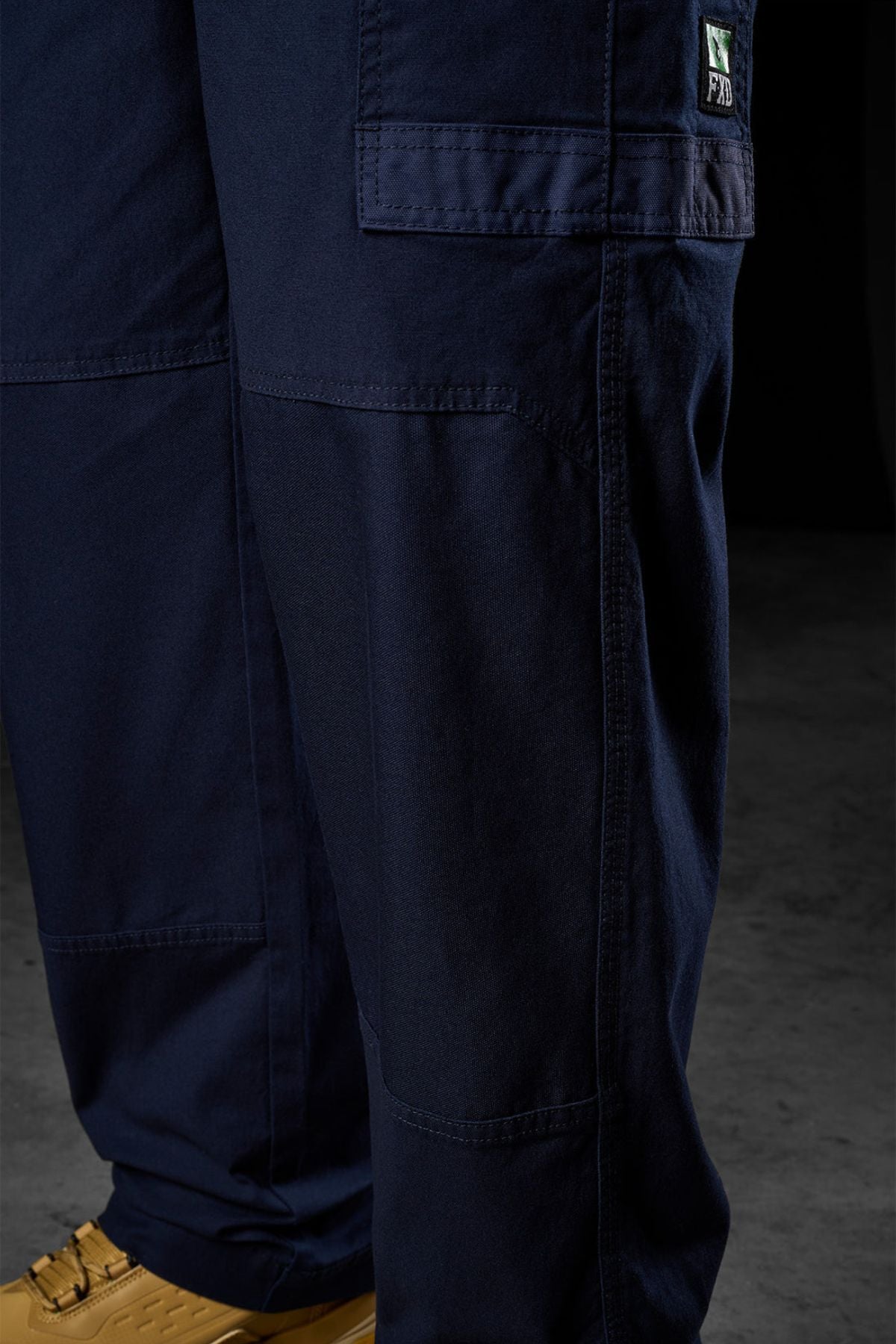 Close-up of a person wearing FXD WP-3L Loose Fit Stretch Canvas Work Pants in navy blue, featuring visible pockets and stitching. Only the lower legs and tan shoes are visible as they stand on a concrete floor.