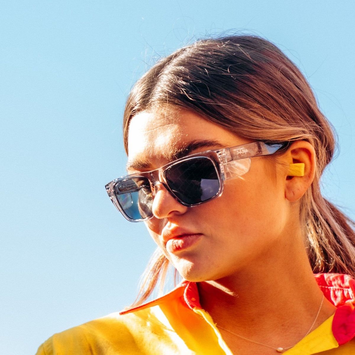 A woman with long hair wears large SafeStyle Fusions Graphite Frame Polarised Lens sunglasses and a colorful shirt in yellow and red hues. She gazes sideways against a clear blue sky, earplugs in place, relishing enhanced clarity without glare.