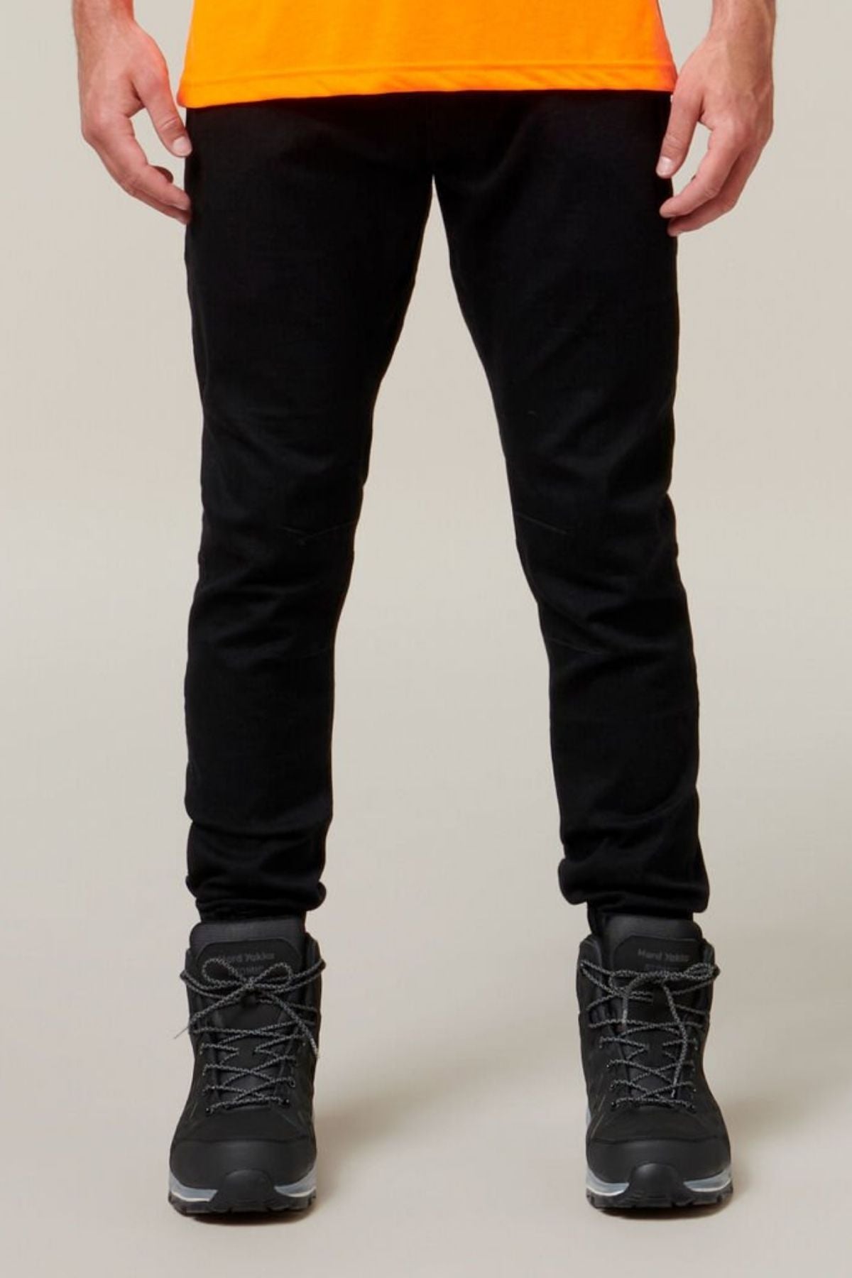 A person wearing Hard Yakka Active Denim Jogger Pant with an elastic waist, black hiking boots with grey laces, and an orange top is photographed from the waist down against a neutral background.