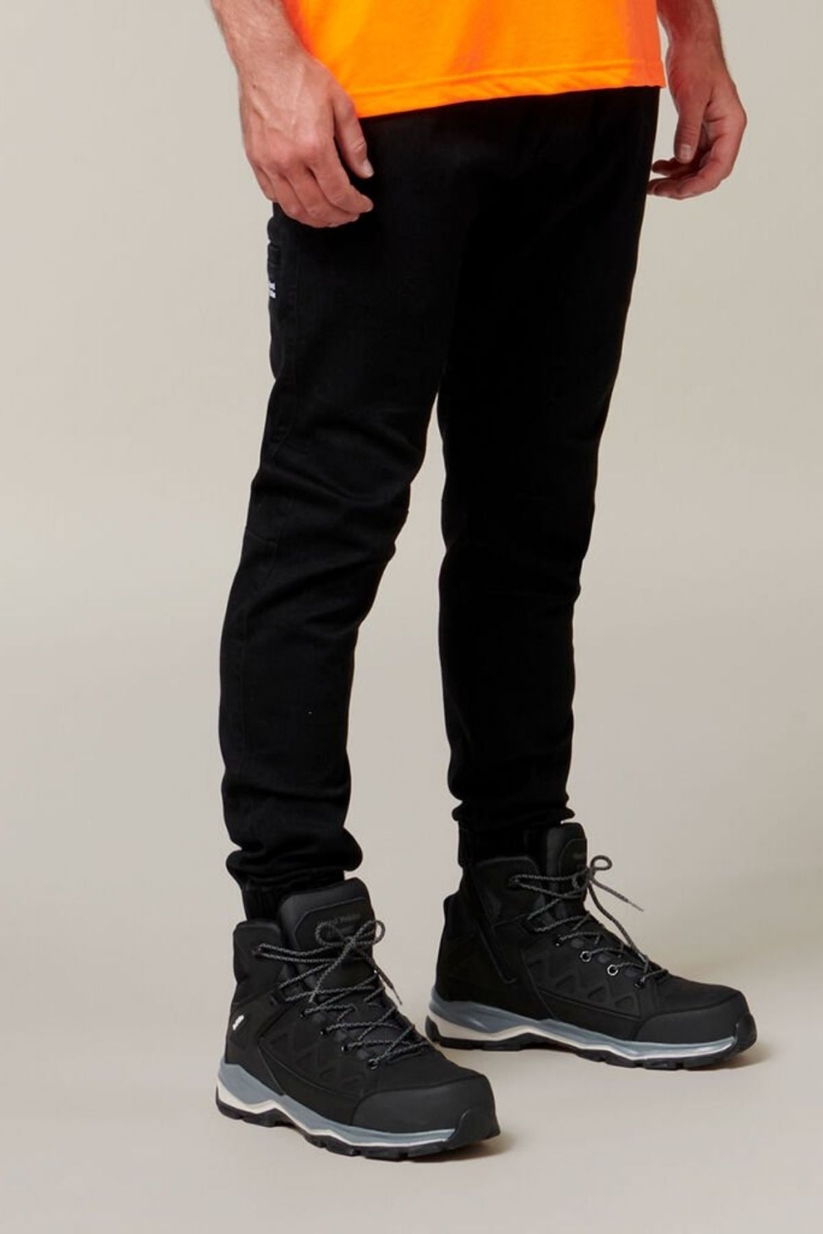A person wearing a bright orange shirt, Hard Yakka Active Denim Jogger Pants with an elastic waist, and black high-top sneakers with grey soles and grey laces stands against a neutral background. The image focuses on the lower half of the body.