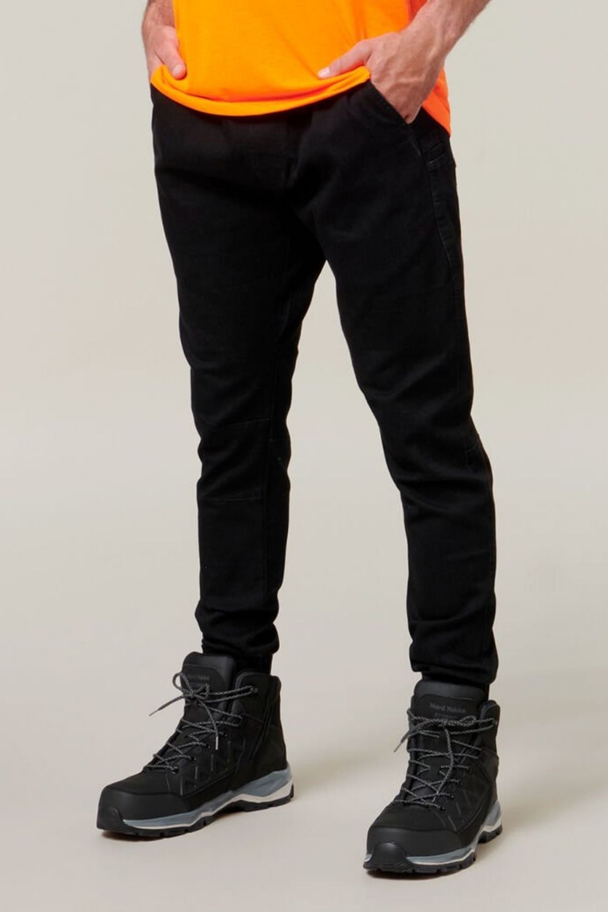 A person wearing Hard Yakka Active Denim Jogger Pants with an elastic waist and black hiking boots stands with their hands in their cargo pockets. The individual is also wearing a bright orange shirt. The background is plain and light-colored.