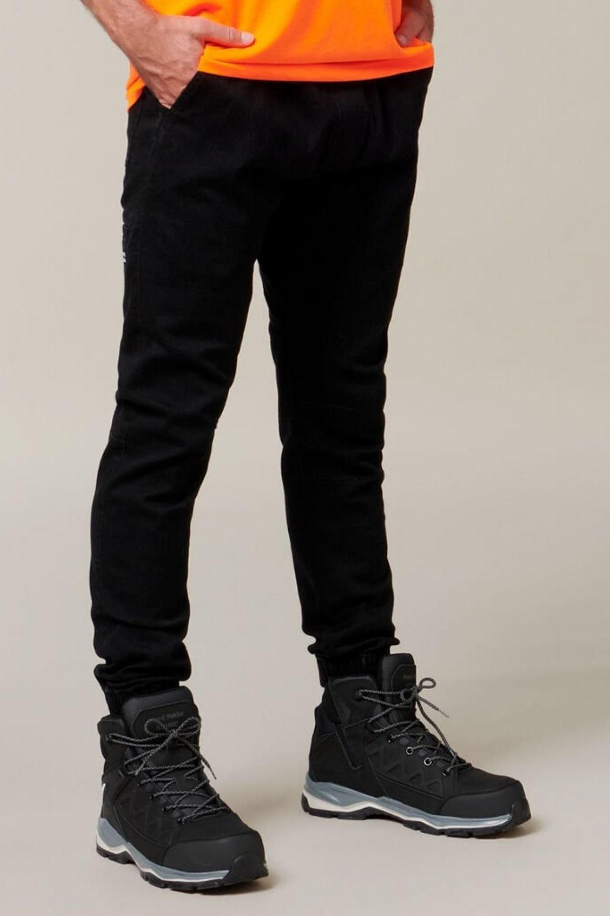 A person is wearing the Hard Yakka Active Denim Jogger Pant with an elastic waist, paired with black hiking boots featuring gray soles. They are seen from the waist down, with their hands in their pockets. The individual is also wearing an orange shirt against a plain, light-colored background.