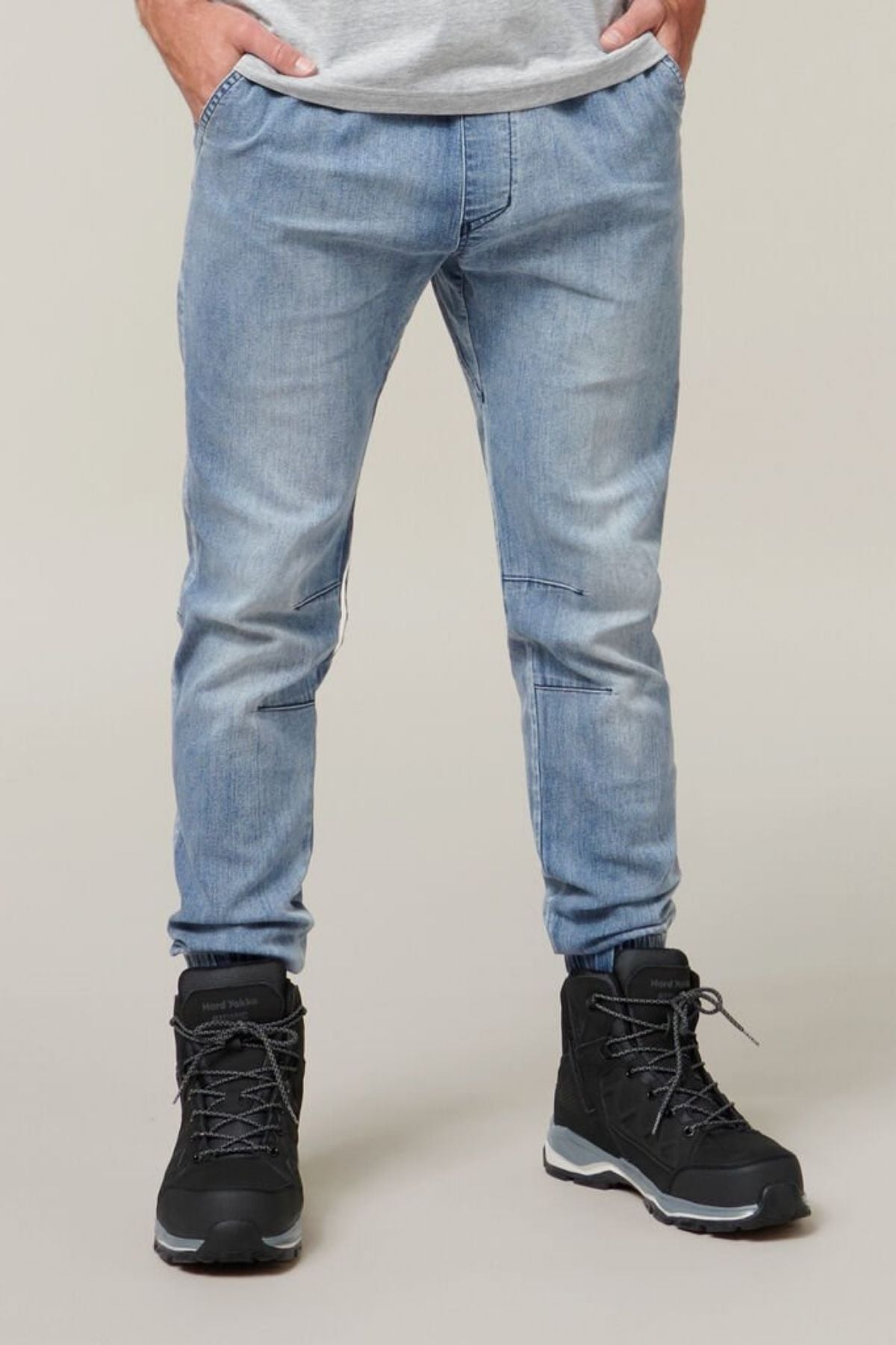 A person wearing Hard Yakka Active Denim Jogger Pants in light blue with cargo pockets and black hiking boots stands with hands in pockets. The background is a neutral color. The person's upper body is partially visible, dressed in a light-colored shirt, exuding a vibe of comfortable wear.
