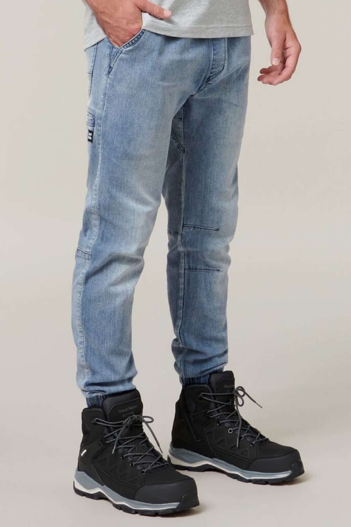 A person is shown from the waist down, wearing Hard Yakka Active Denim Jogger Pants in light blue and black hiking boots with gray accents and laces. One hand is in their pocket, exuding a sense of comfortable wear, and the background is plain and neutral.