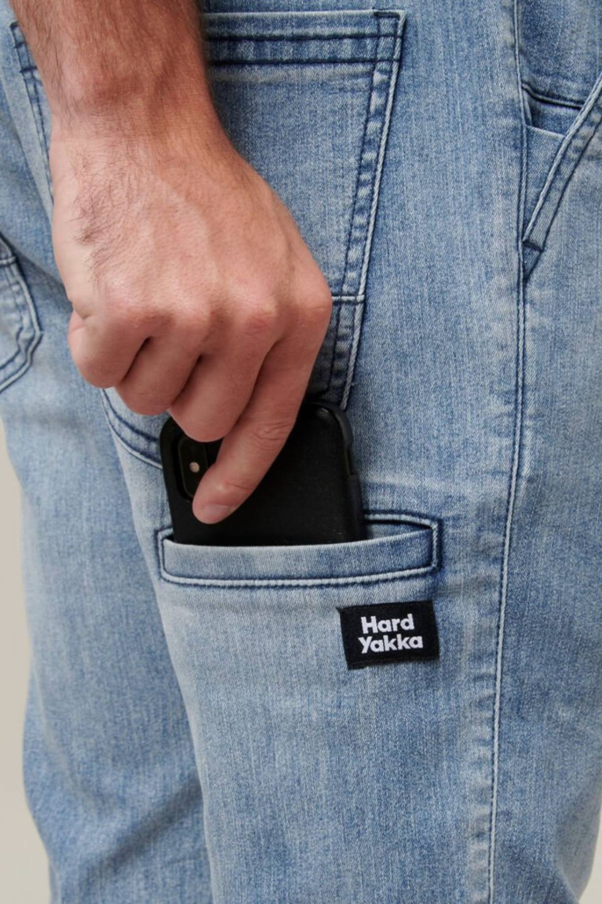 A person wearing Hard Yakka Active Denim Jogger Pants is shown placing a black smartphone into a side pocket. Only the person's hand and lower torso are visible, showcasing the practical and comfortable wear of these sturdy trousers.