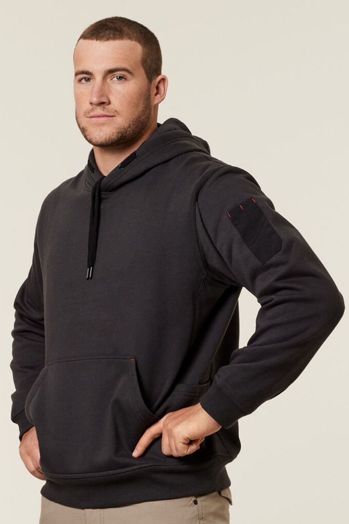 A man with short hair and a trimmed beard wears the Hard Yakka Plain Fleece Hoodie in black, featuring kangaroo pouch pockets and a sleeve patch, standing with one hand on his hip against a plain light background.