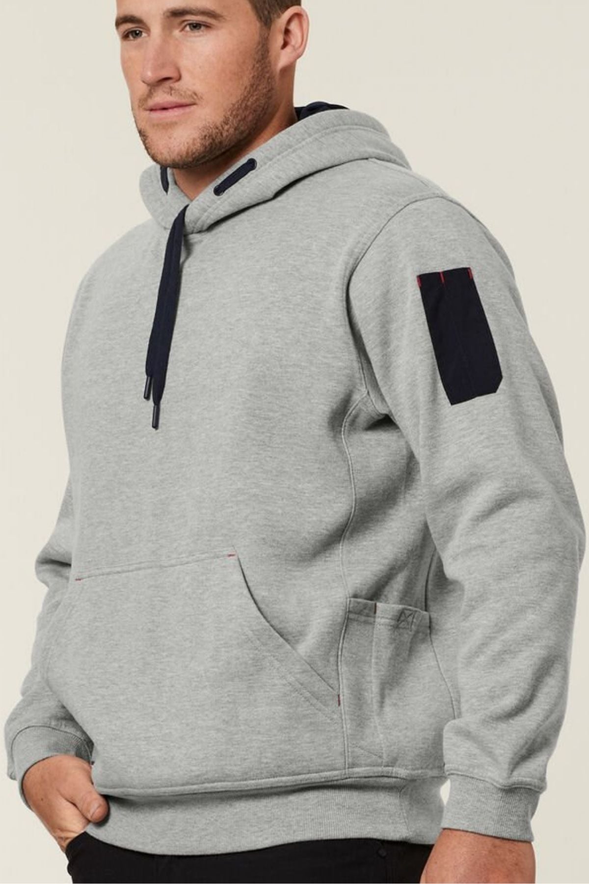 A man with light skin and short hair wears a Hard Yakka Plain Fleece Hoodie made from brushed fleece, featuring a dark drawstring and a black rectangular patch on the upper sleeve, standing against a plain beige background.