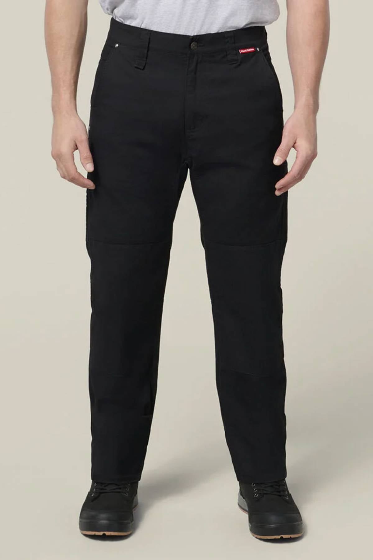 A person wearing Hard Yakka Heritage Carpenters Pant in black, a light gray t-shirt, and black shoes stands facing forward with arms relaxed by their sides against a plain beige background.