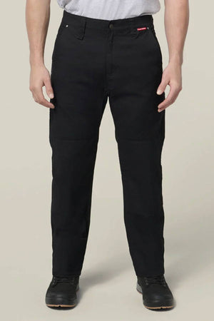 A person wearing Hard Yakka Heritage Carpenters Pant in black, a light gray t-shirt, and black shoes stands facing forward with arms relaxed by their sides against a plain beige background.