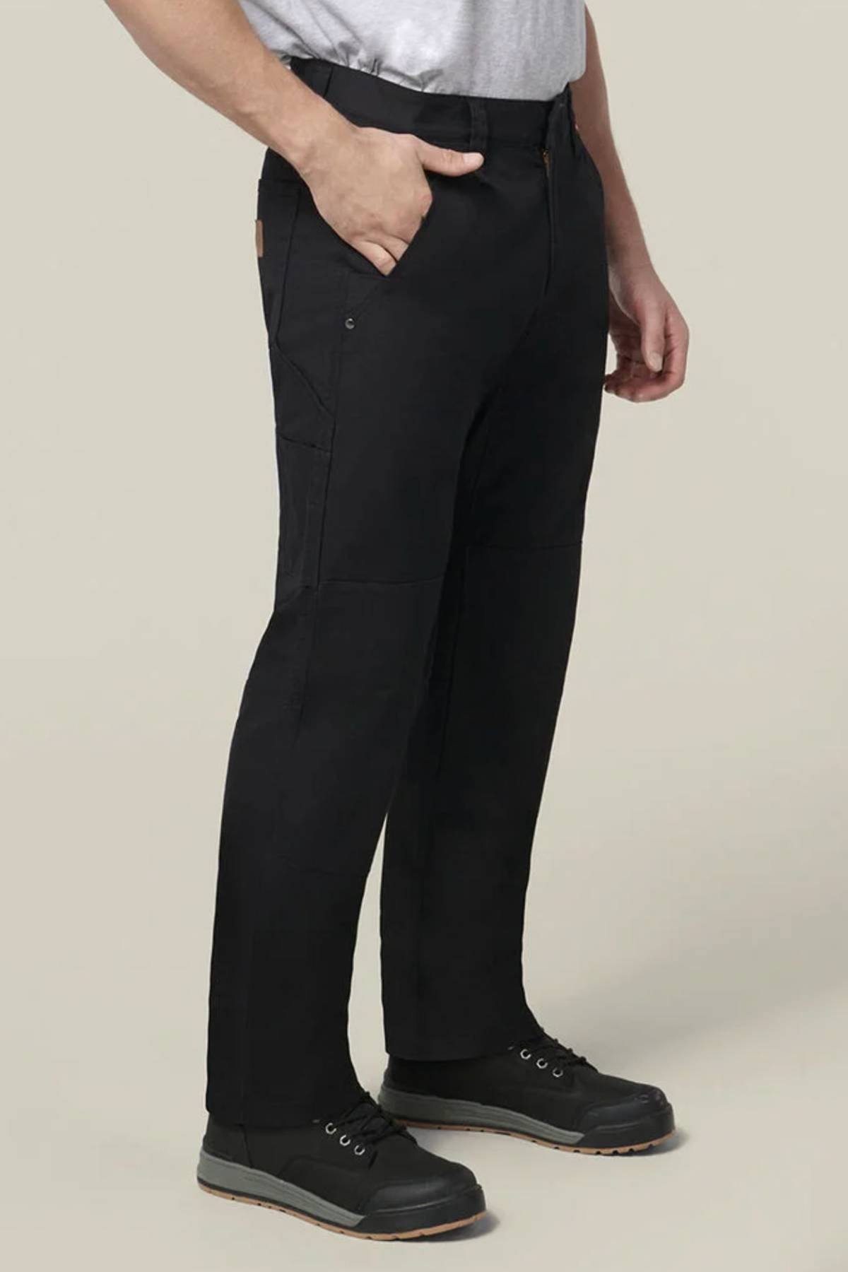 A person wearing Hard Yakka Heritage Carpenters Pant in black, a light gray t-shirt, and black work shoes stands with one hand in their pocket against a plain light background.
