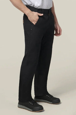 A person wearing Hard Yakka Heritage Carpenters Pant in black, a light gray t-shirt, and black work shoes stands with one hand in their pocket against a plain light background.